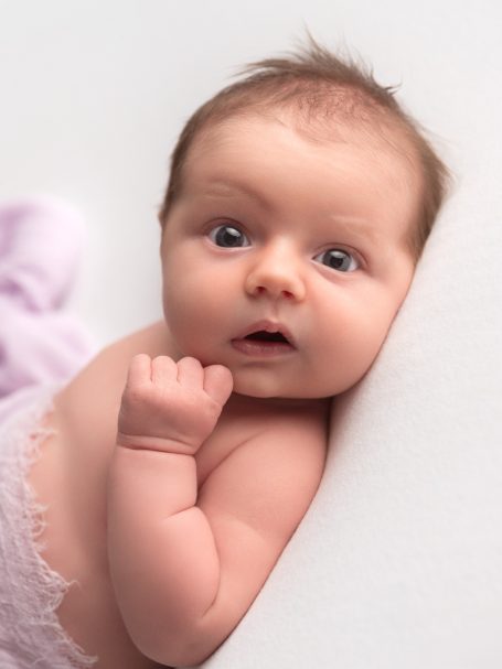 Newborn baby with wide eyes, resting on a soft surface, looking curiously at the camera.